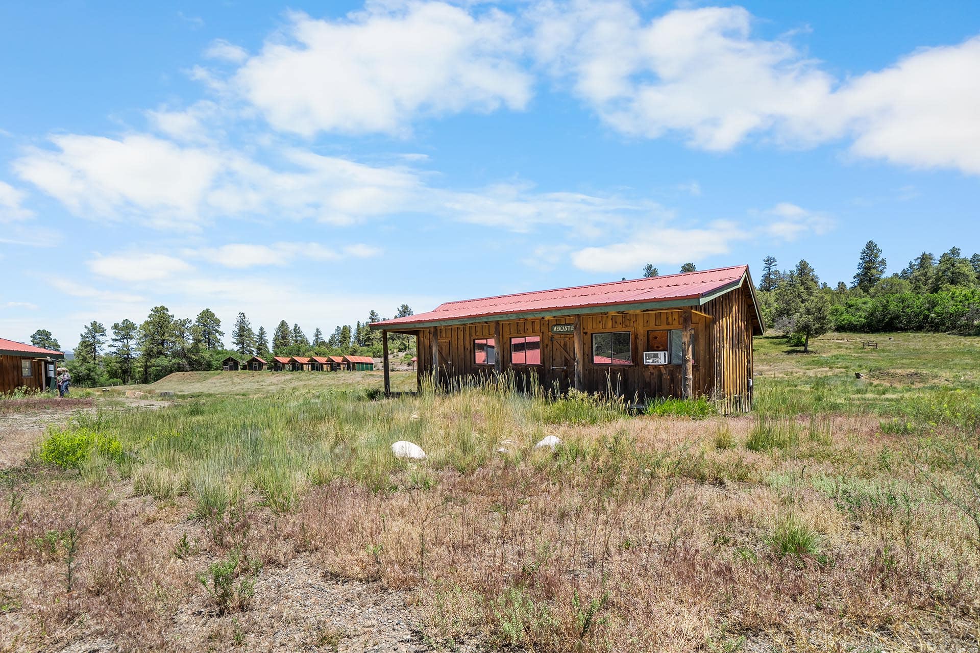 Rustic wooden building with a red metal roof in an open grassy field under a bright blue sky with scattered clouds.