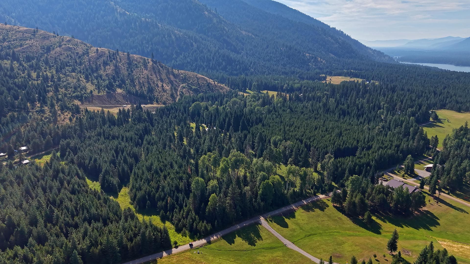 Aerial view of a forested mountain valley with winding roads and distant lake on the horizon.