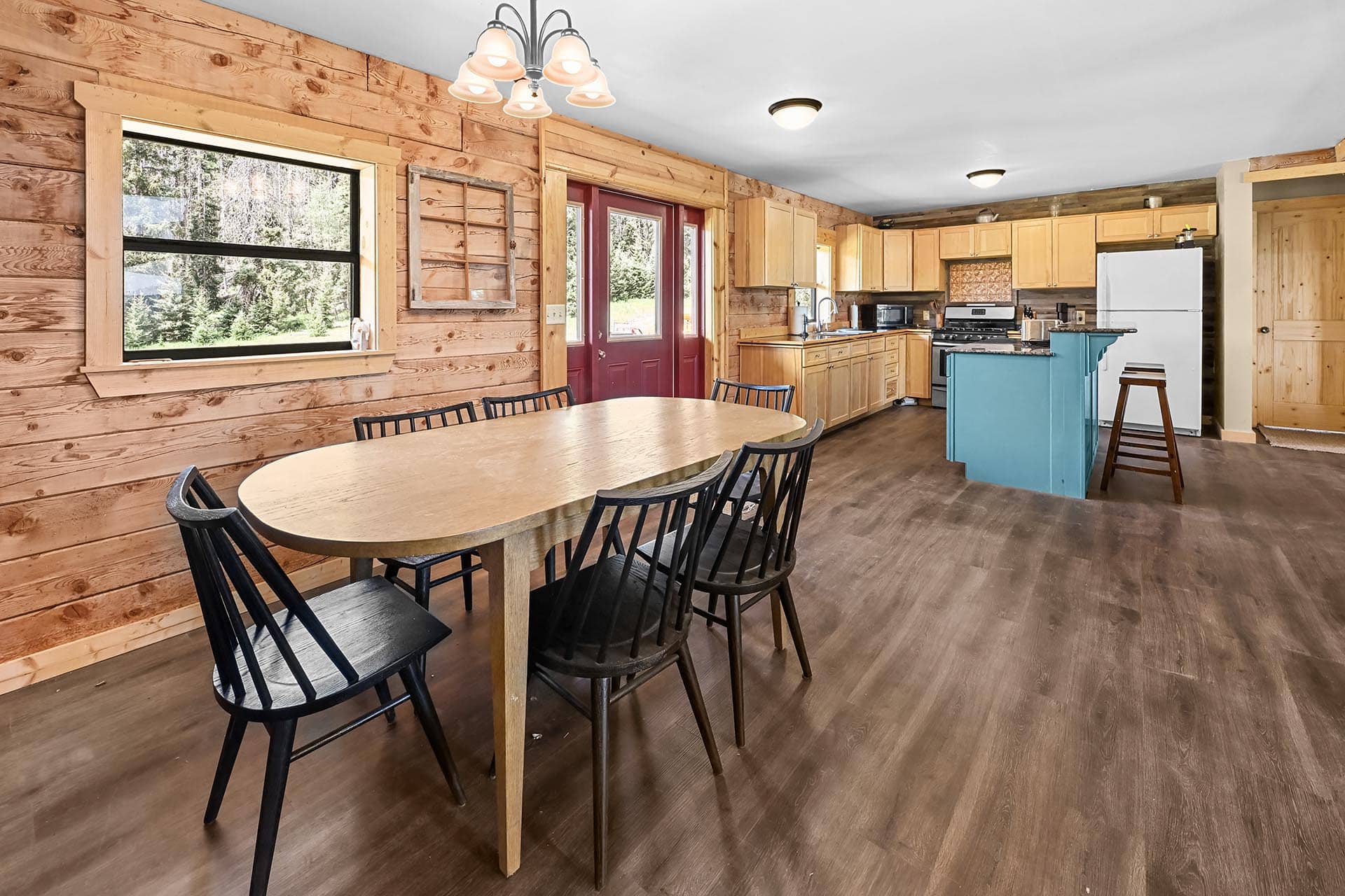 Rustic cabin kitchen and dining area with an oval wooden table, black chairs, and a teal kitchen island near a red entry door.
