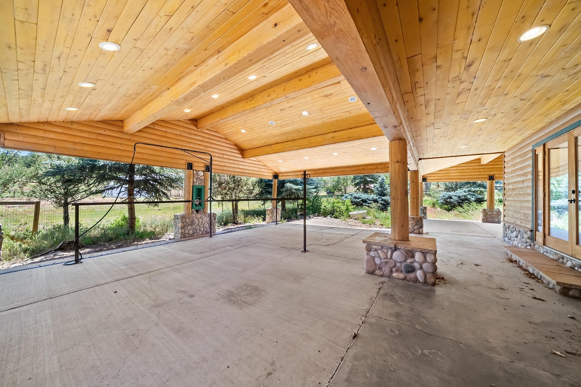 Open-air wooden pavilion with stone column bases and recessed ceiling lights, overlooking a garden area