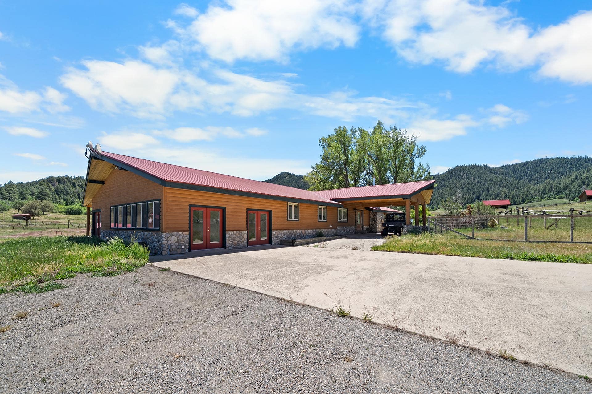 Single-story wooden building with a red metal roof in a rural setting, mountains in the background under a blue sky with clouds.