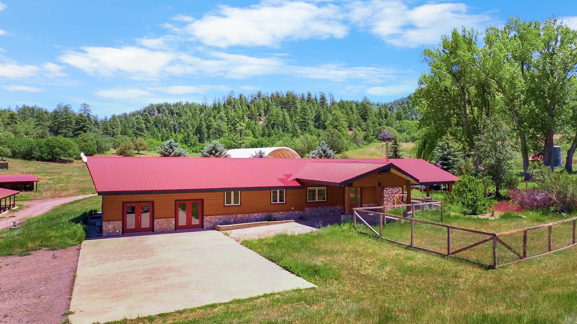 Single-story wooden house with a red metal roof and stone foundation, set in a green rural yard with trees and hills in the background.