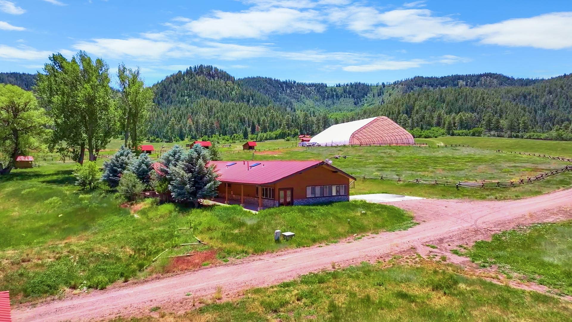 Rural farm with a red-roofed building, a large white greenhouse, and green fields backing a forested hillside under a blue sky.