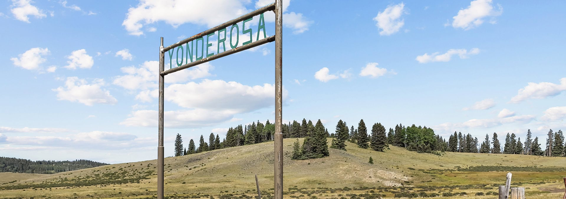 Rusty metal roadside sign reading 'Yonderosa' against a blue sky and grassy hill with a line of pines nearby.