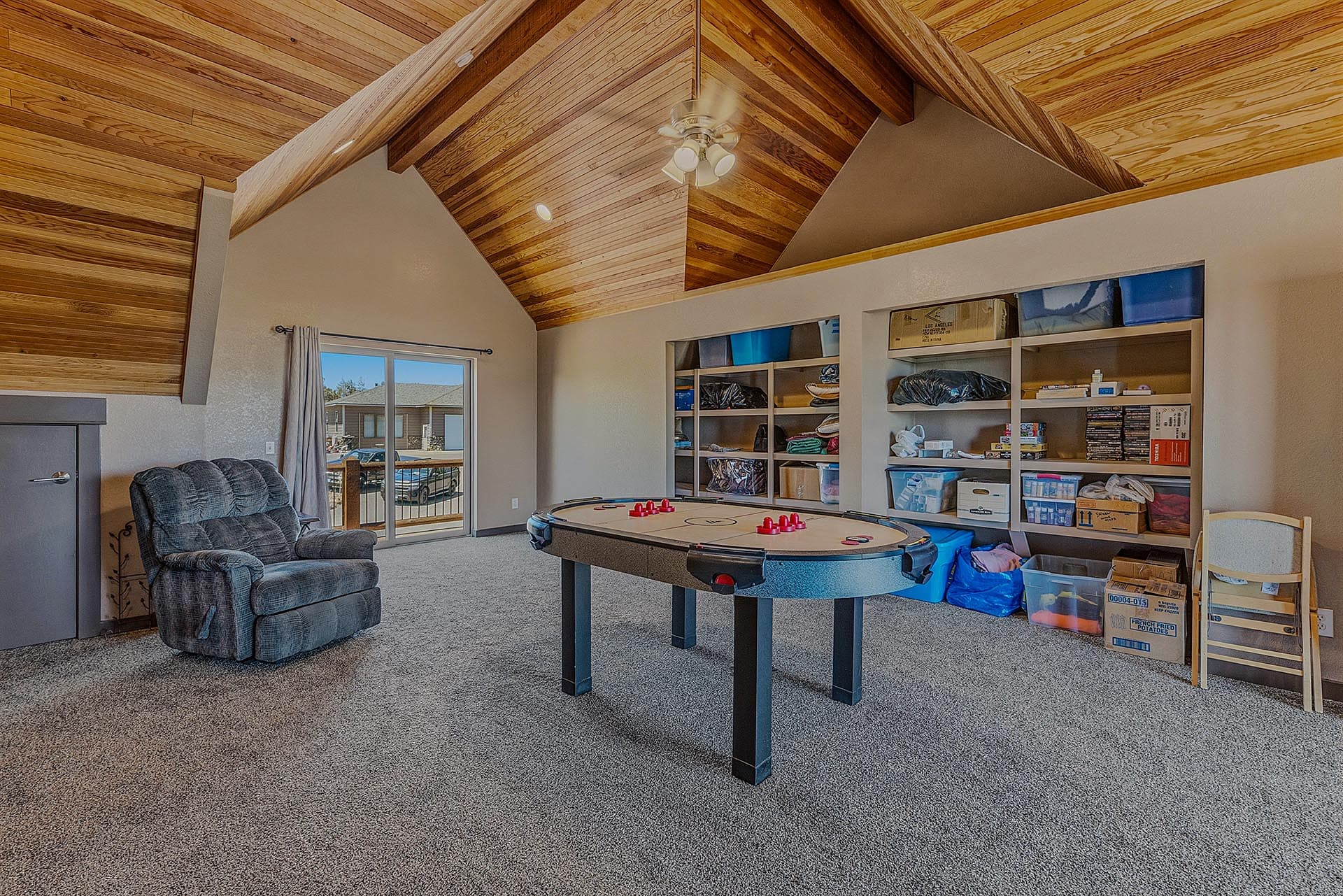 Recreational game room with an air hockey table, stacked shelves of storage boxes, and a recliner chair under a wooden vaulted ceiling.