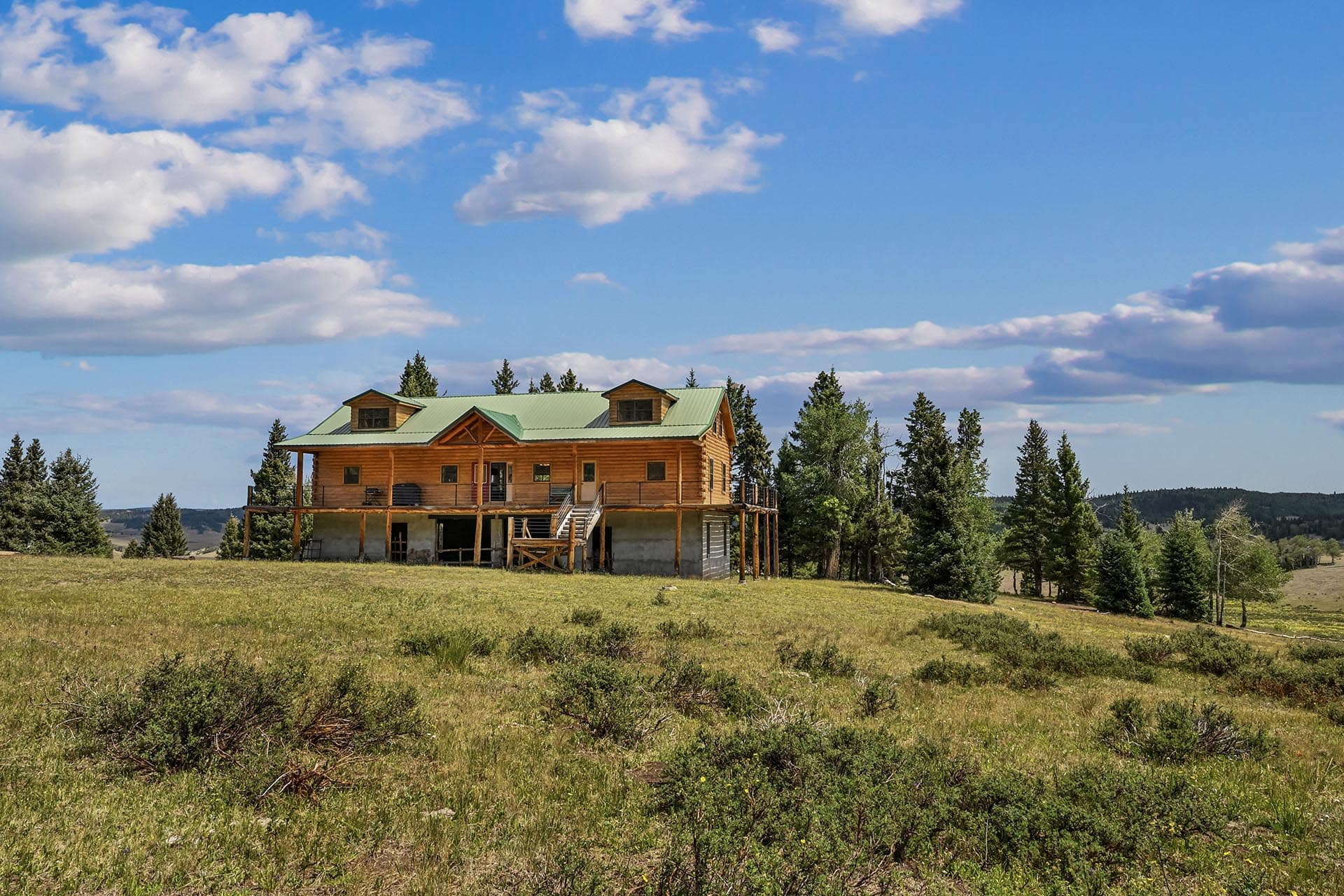 Wooden cabin on stilts under construction in a grassy field, with pine trees and a bright blue sky.