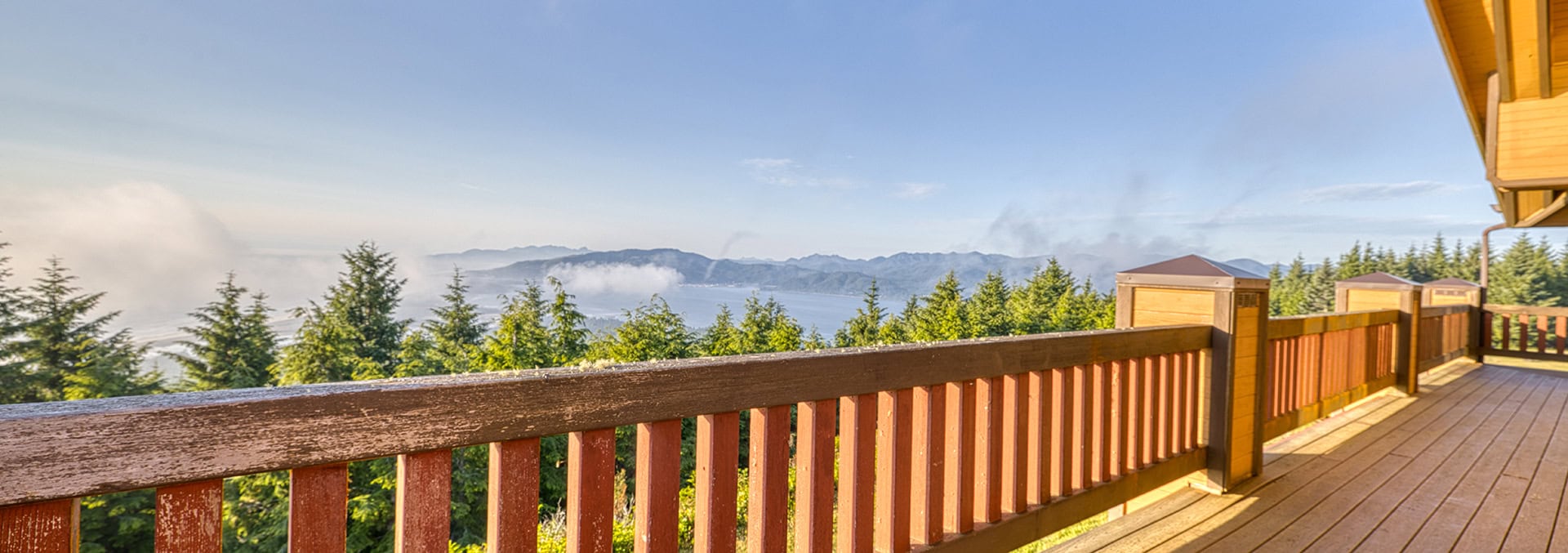 Wooden deck railing with a forest and distant mountain range under a clear blue sky, sunny day view from a balcony.