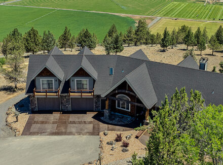 Aerial view of a large dark-roofed house with garages, surrounded by trees on a rural property and green fields beyond.