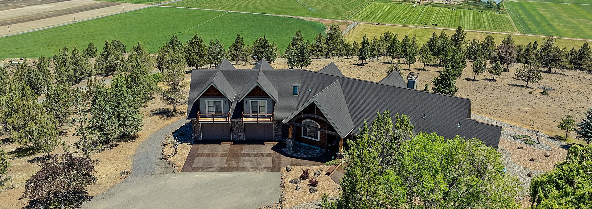 Aerial view of a large dark-roofed house with garages, surrounded by trees on a rural property and green fields beyond.