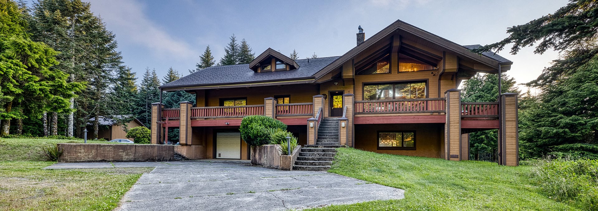 Wood-and-stone two-story house with a large wraparound balcony set among tall evergreen trees.