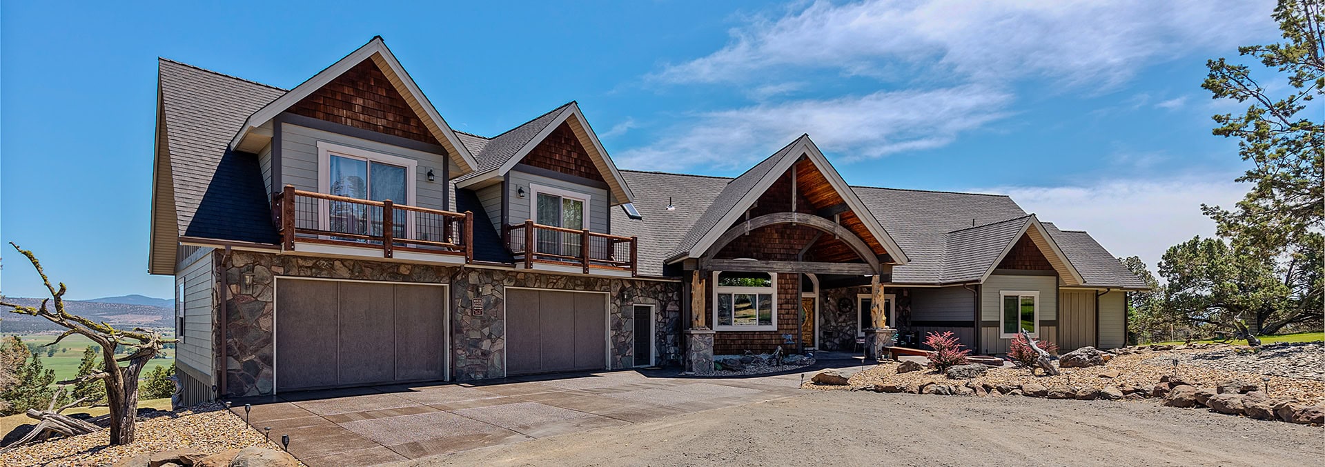 Two-story house with stone foundation and wood siding, multiple gables, balconies, and a covered front entry under a bright blue sky.