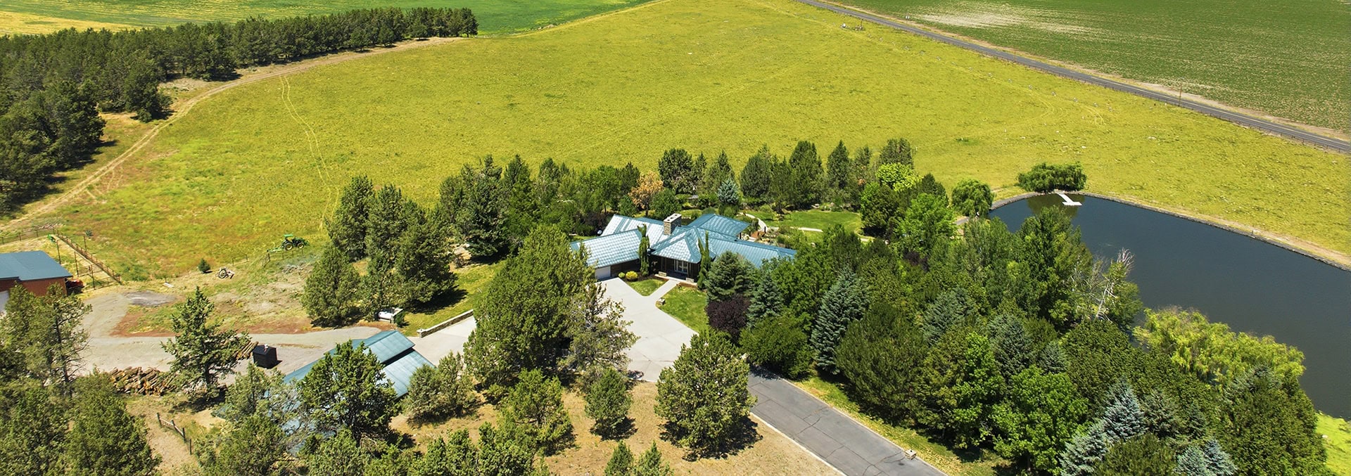 Aerial view of a rural property with a blue-roofed house, driveway, and dense surrounding trees near a curved pond far to the right.