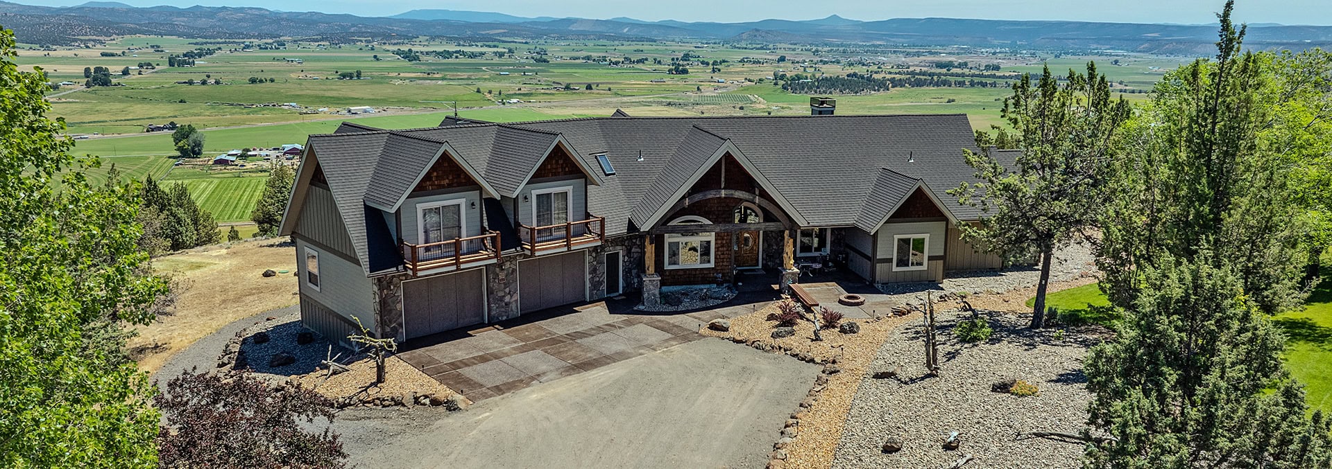 A large modern house with a dark gray roof, stone and wood siding, and a three-car garage, set in a dry landscaped yard with gravel and trees; rural valley beyond.