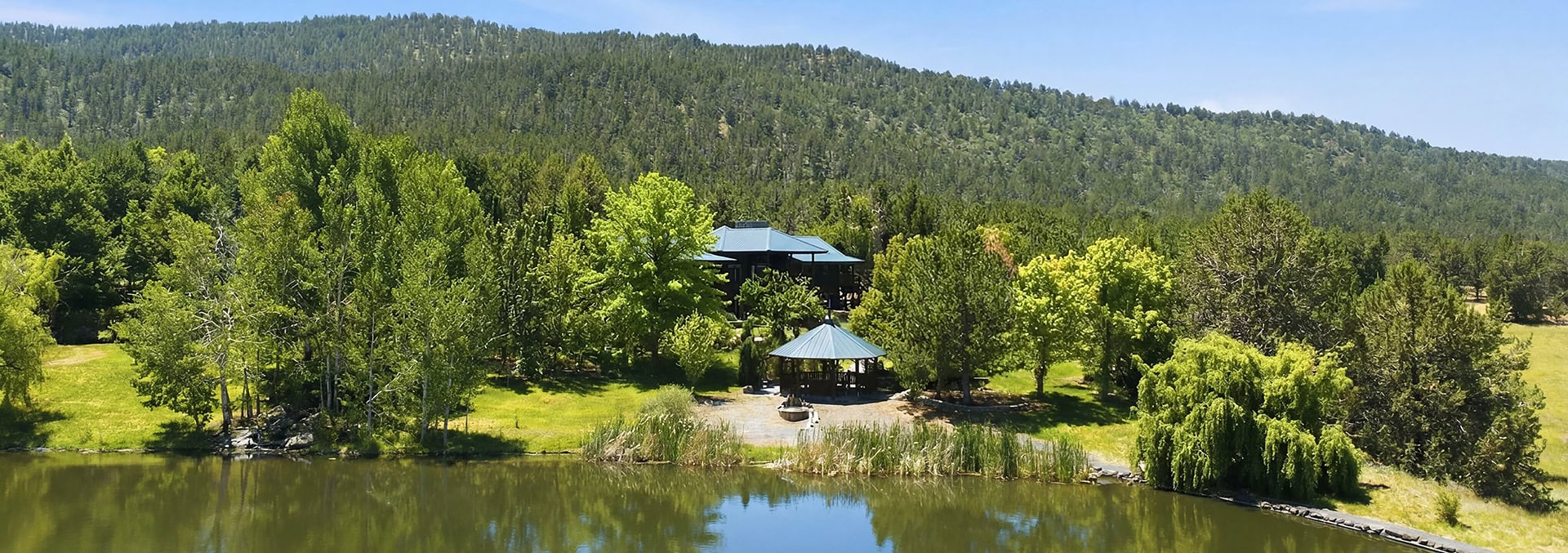 Lakeside park with a blue-roofed pavilion and a gazebo among green trees, hills in the background.