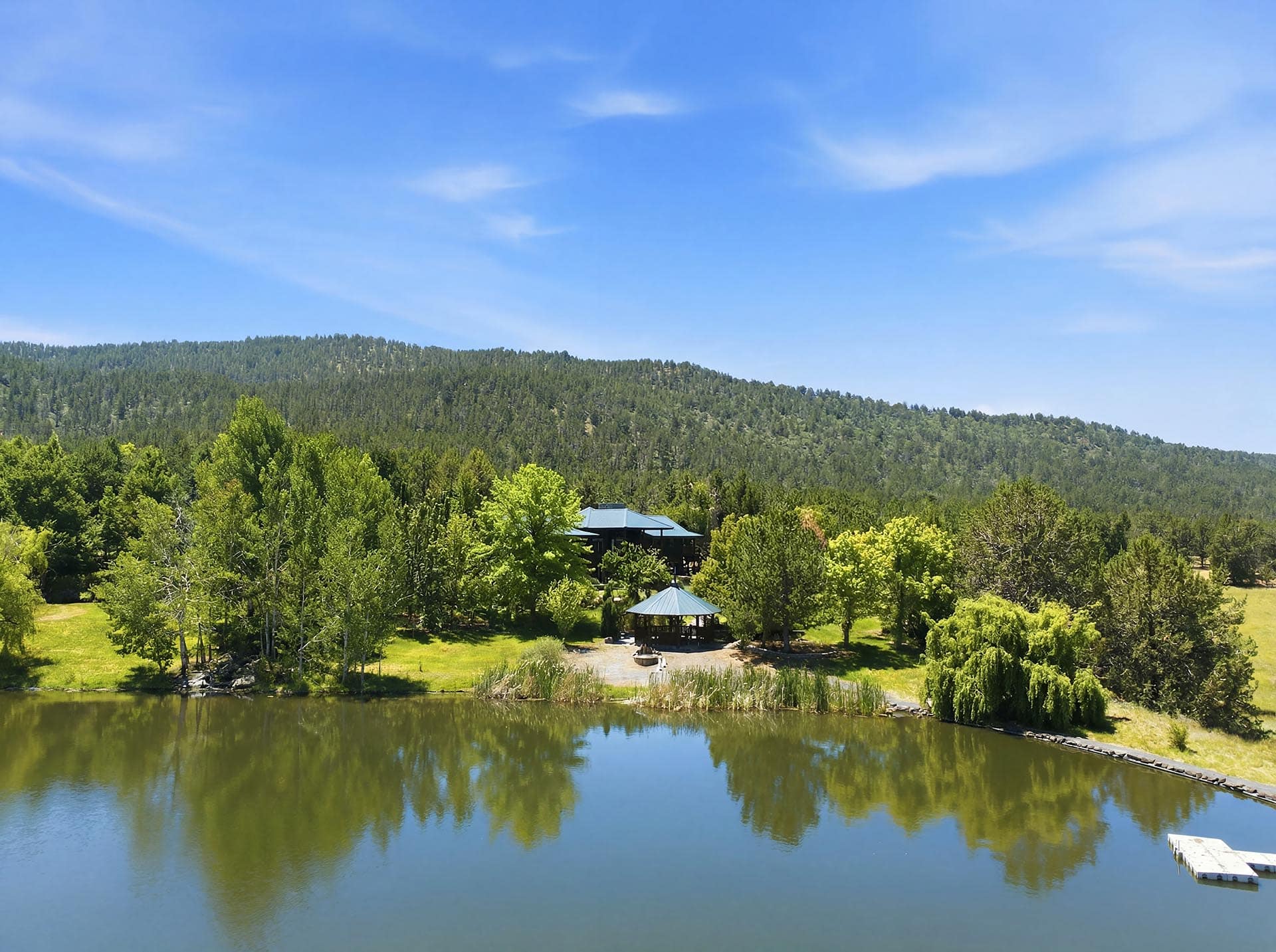 A calm lake edged by green trees with a pavilion and blue-roofed structures among the shore foliage, hills in the background under a clear blue sky