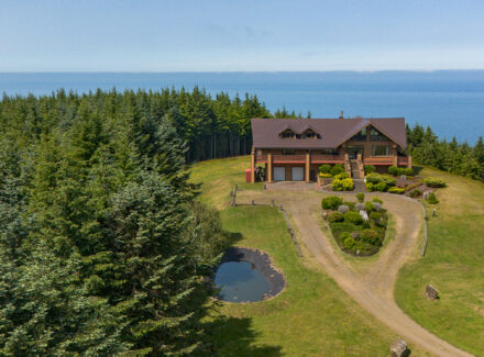 Chalet-style house on a grassy hill with a curved driveway, surrounded by evergreen trees and a small pond, overlooking the ocean.