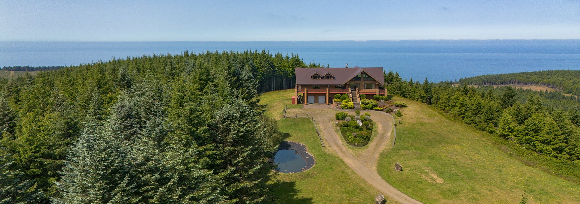 Chalet-style house on a grassy hill with a curved driveway, surrounded by evergreen trees and a small pond, overlooking the ocean.