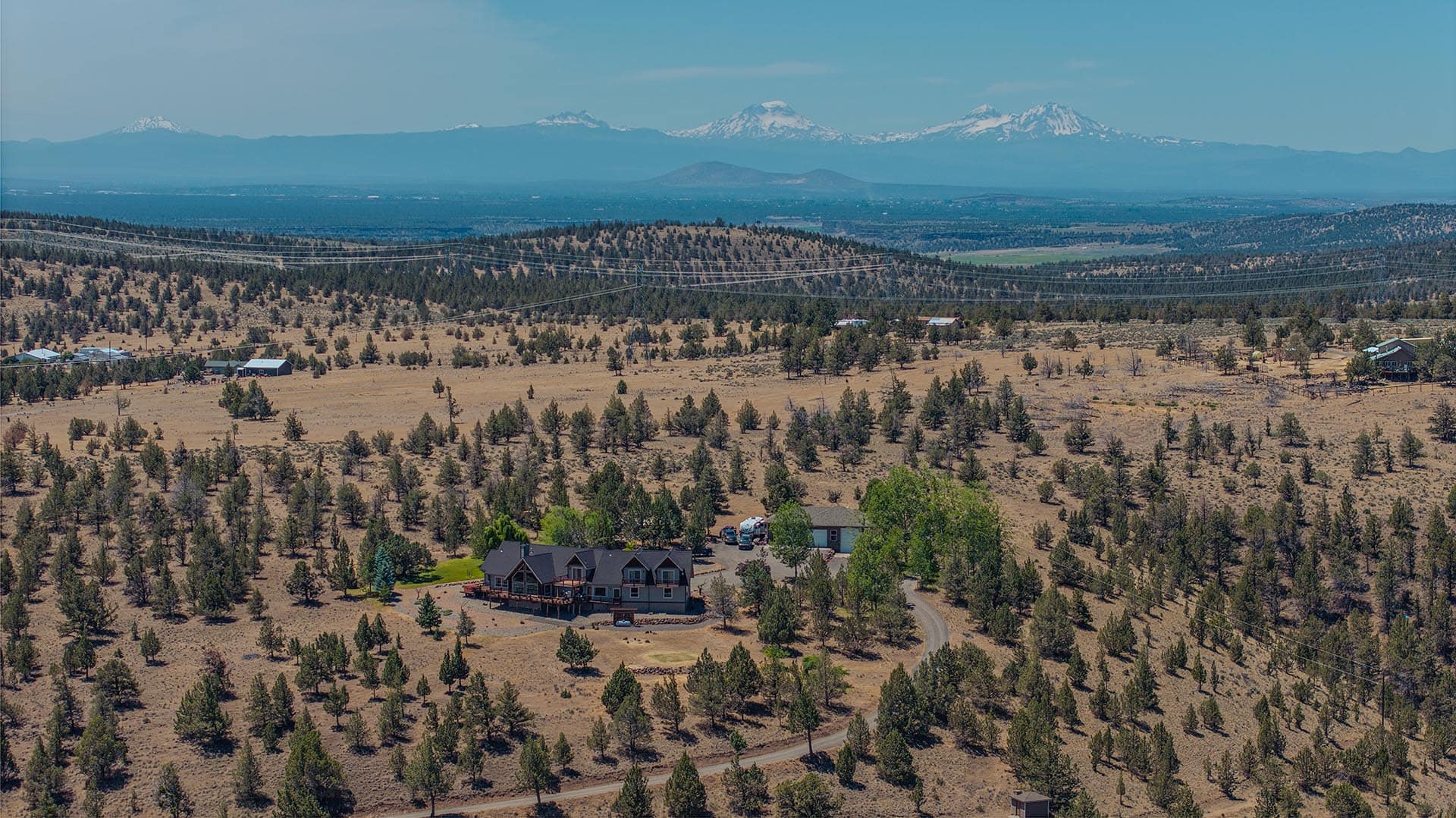 Rural landscape with a large house in the foreground, scattered trees, power lines, and snow-capped mountains on the horizon.