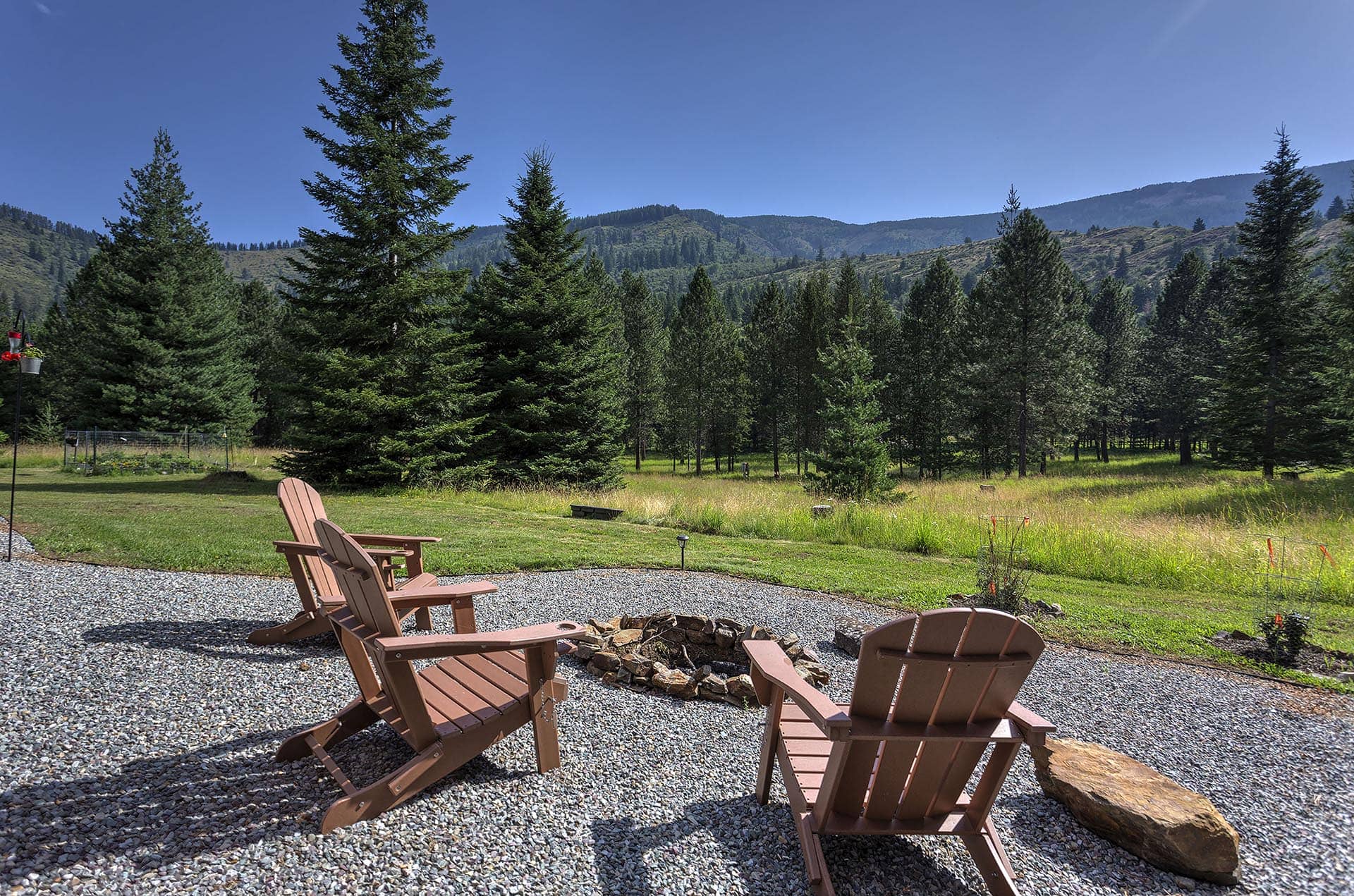 Adirondack chairs around a rock fire pit on gravel, with a grassy yard and pine forest beyond under a clear blue sky.