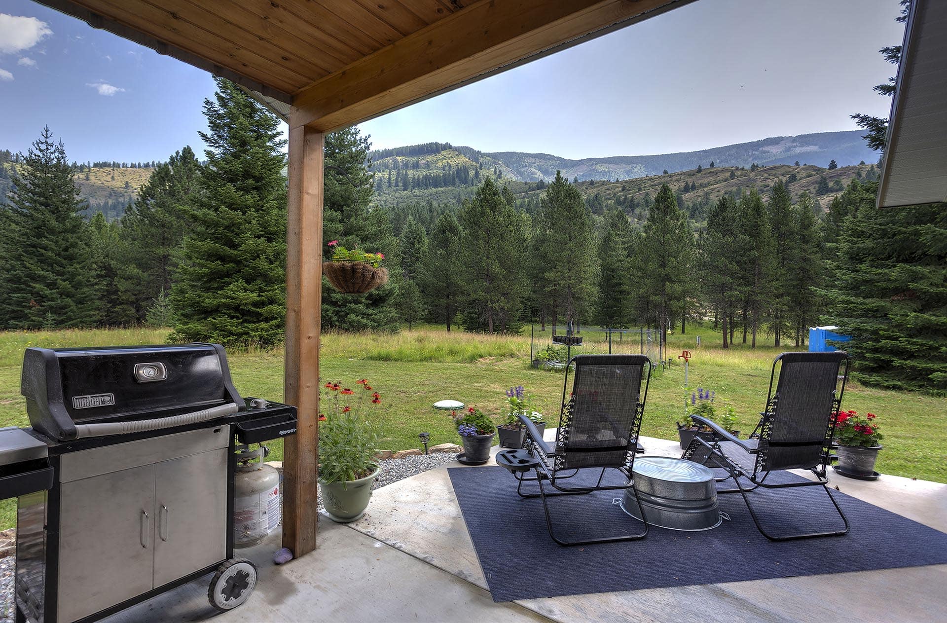 Patio with a grill and four black lawn chairs on a rug, overlooking a forested valley and distant mountains under a clear blue sky, from a covered area.