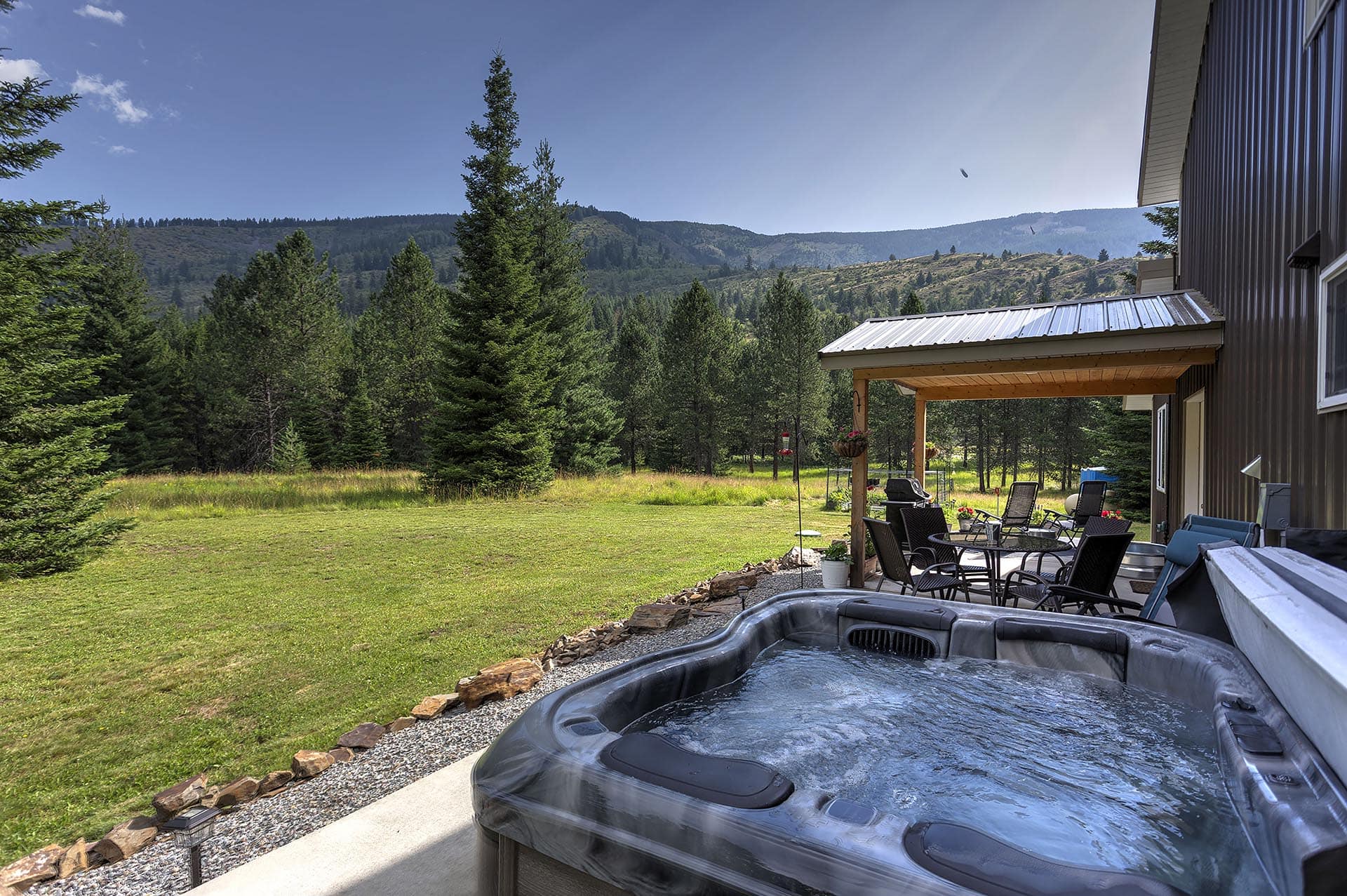 Backyard scene with a dark-siding house, covered patio, and a bubbling hot tub in the foreground, overlooking a forest and mountain range.
