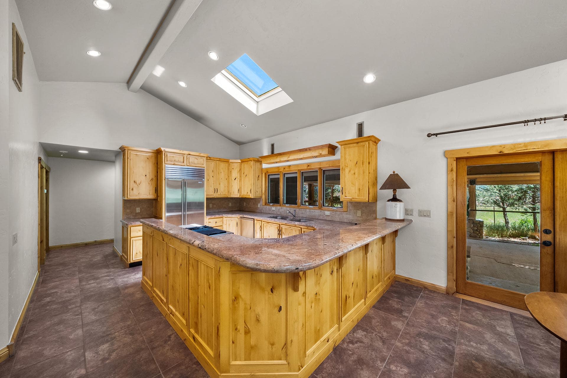 Bright kitchen with natural wood cabinets, a curved granite island, and a skylight overhead. The stainless fridge is visible along the back wall.
