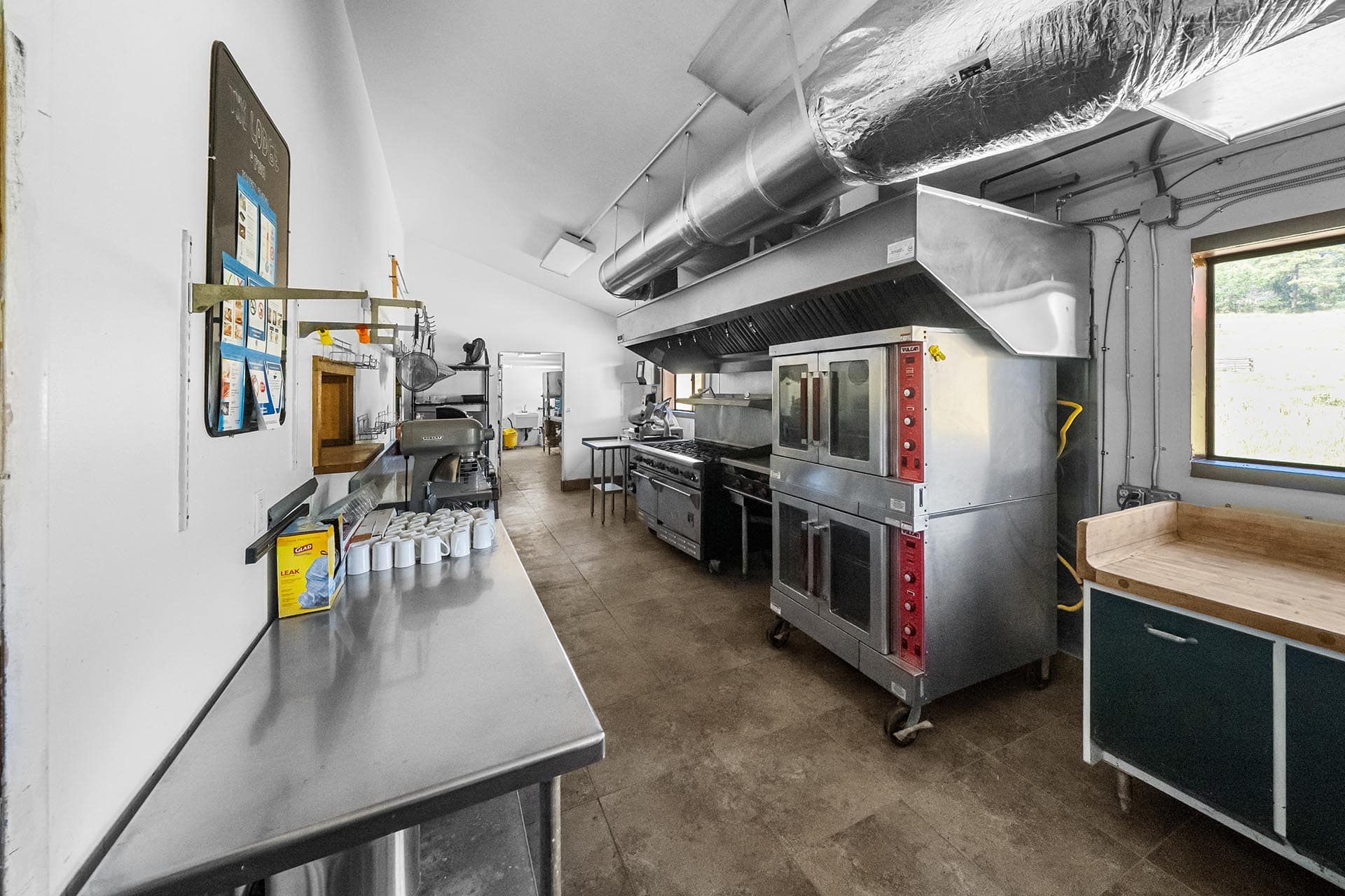 Commercial kitchen interior with stainless steel ovens, vent hood, and prep counter; window to outside on right side.