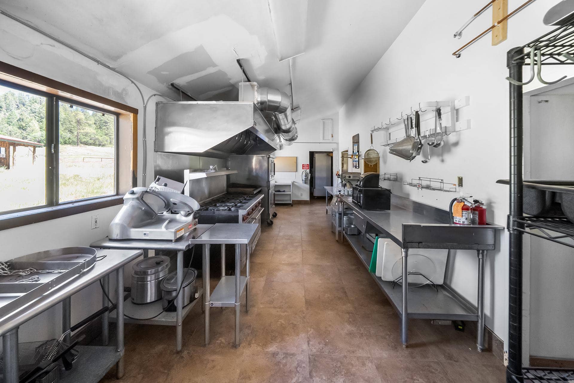 Commercial kitchen with stainless steel appliances, vent hood, and hanging utensils along white walls. Panoramic window to outside on left.