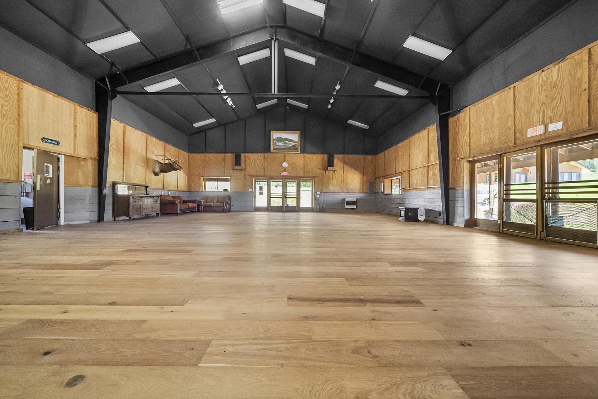 Interior of a large rustic hall with wooden walls, high ceiling, skylights, and empty hardwood floor; deer mount and couches against wall on left.