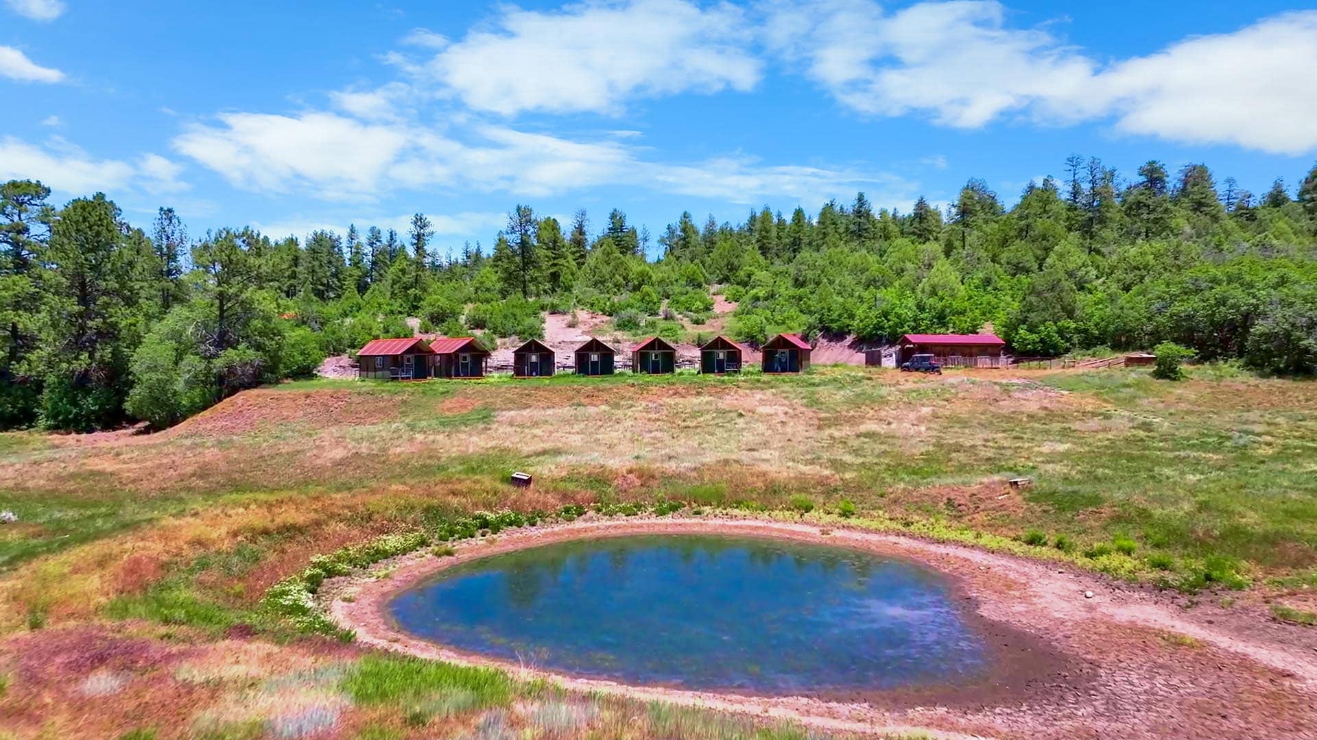 Row of small cabins with red roofs lined along a grassy hillside above a circular pond, with trees and a blue sky in the background.