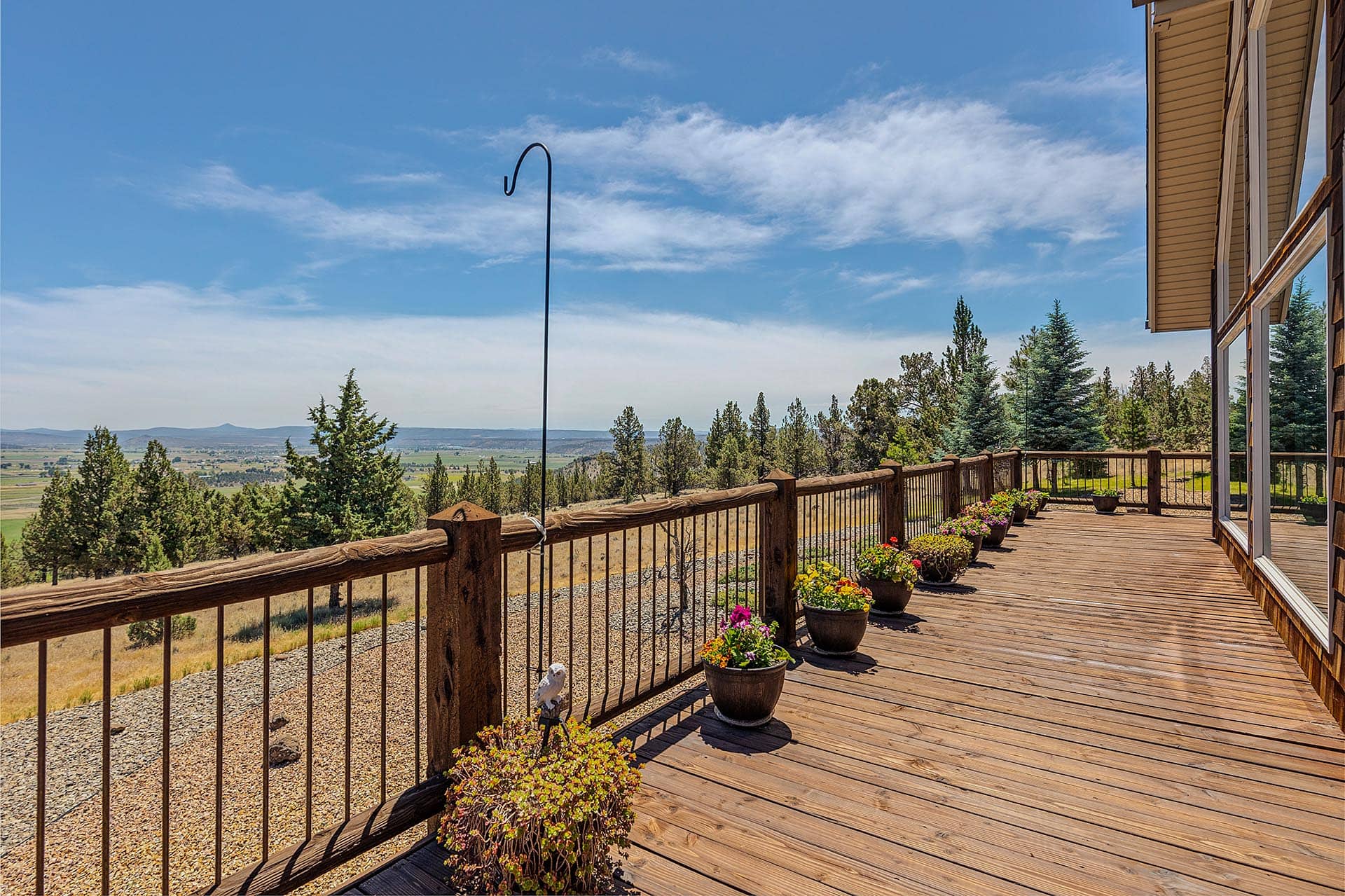Wooden deck with flower pots along a railing, overlooking a valley with pine trees under a blue sky.