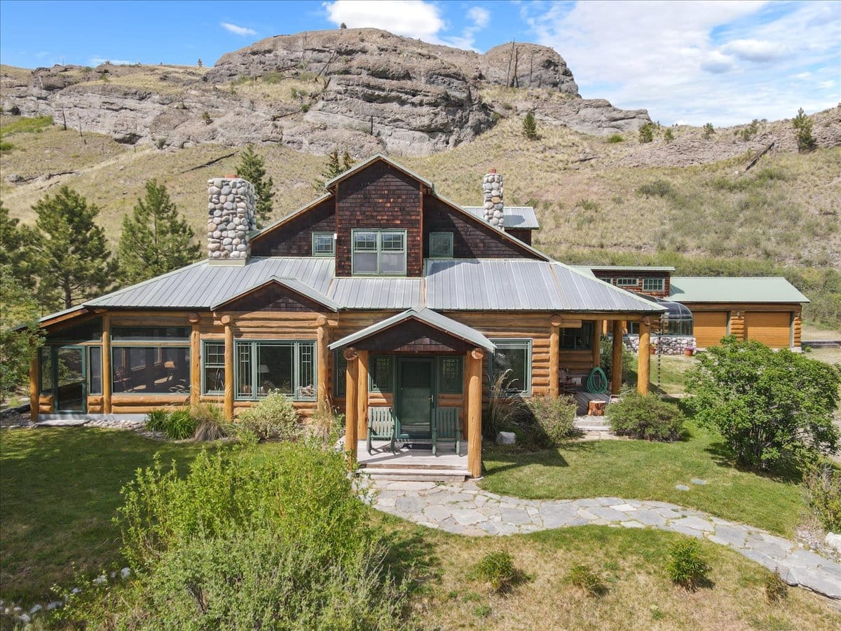Log cabin-style house with a metal roof and stone chimneys seated on a grassy hillside with rocky hills in the background.