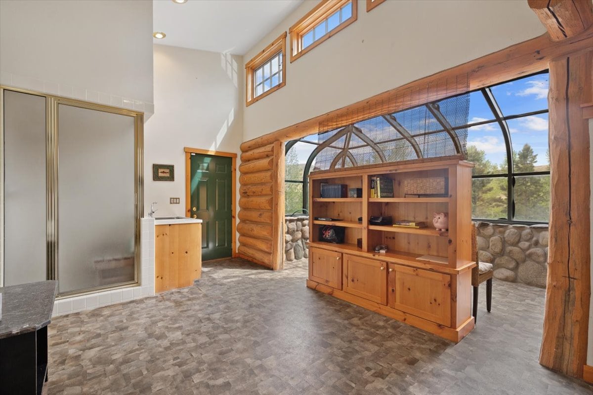 Open-concept rustic living space with log beams, large wooden bookshelf, and curved glass ceiling overlooking stone wall and greenery outside.