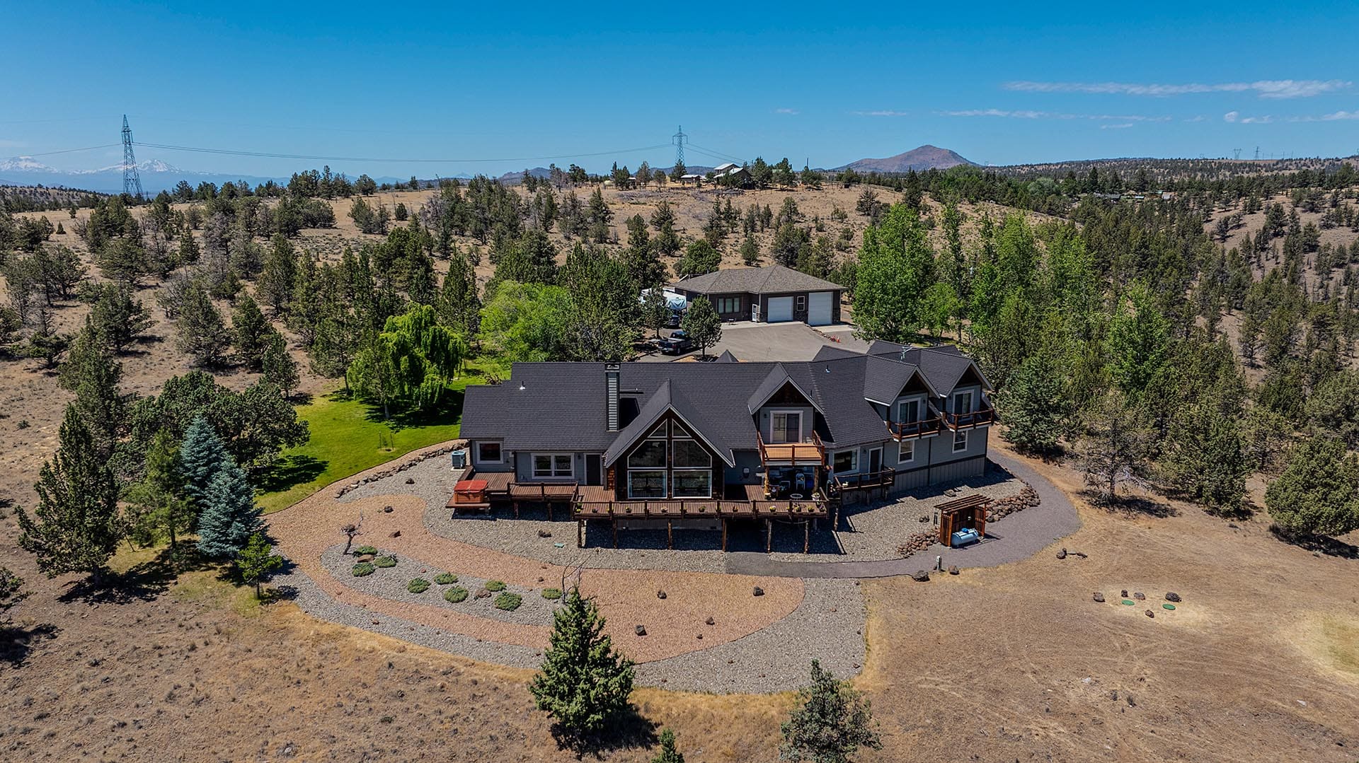 Aerial view of a large two-story house with dark roof and wraparound decks, set on a dryland lot with trees and a curved driveway under a clear blue sky.