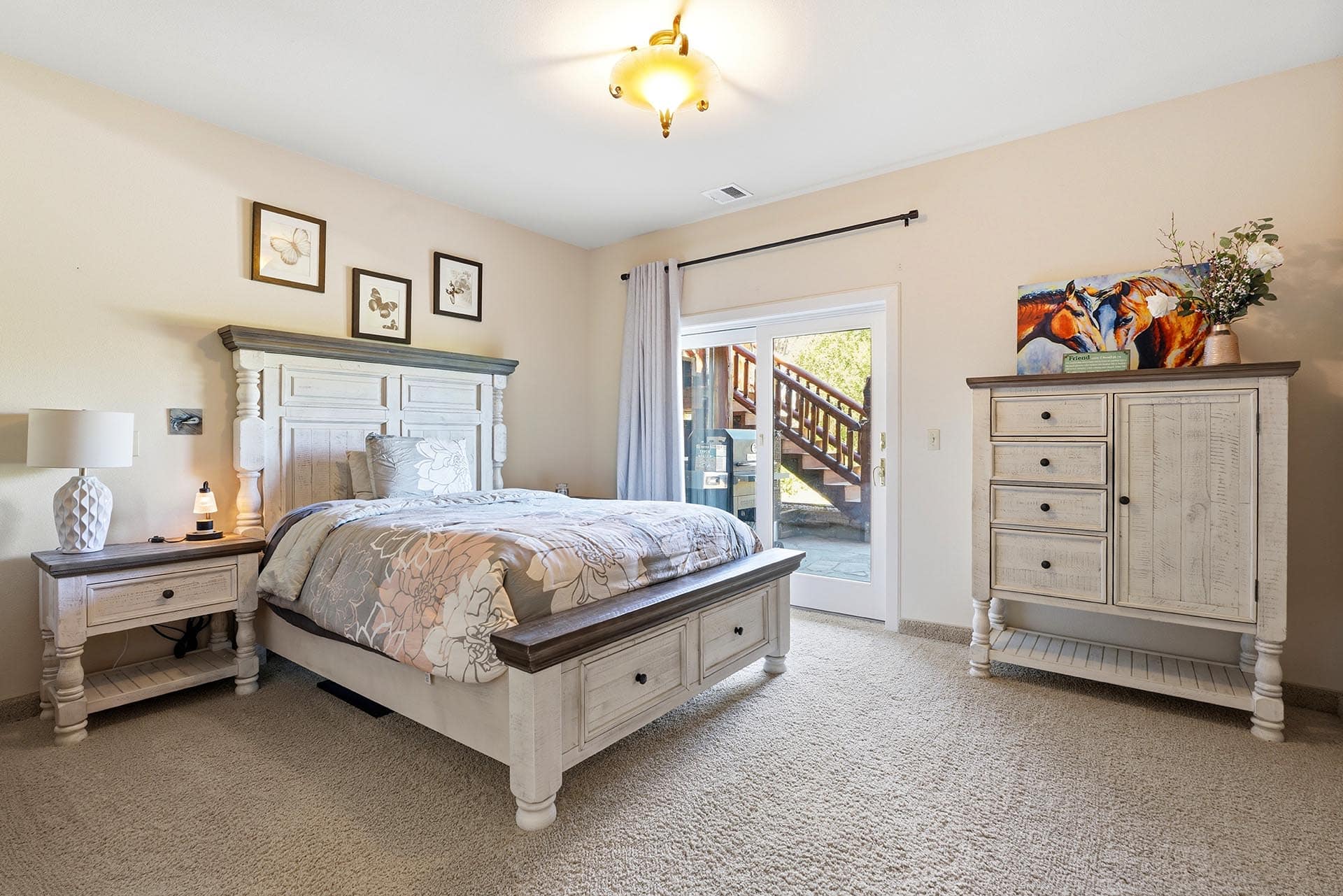 Bedroom with distressed white furniture, floral bedding, beige walls, and a ceiling light fixture; sliding glass door opens to an outdoor stairway.