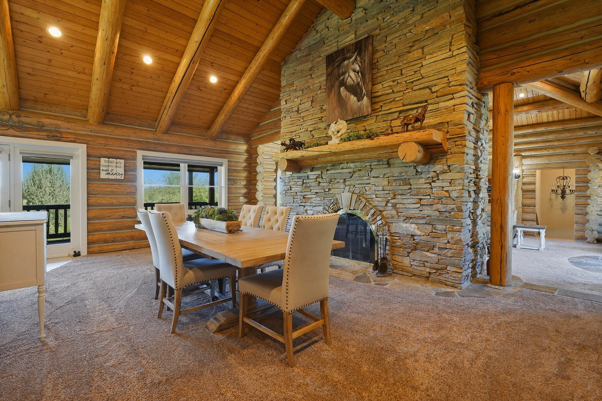 Rustic dining area in a log cabin with a stone fireplace, exposed wooden beams, and a long wooden table with beige upholstered chairs.