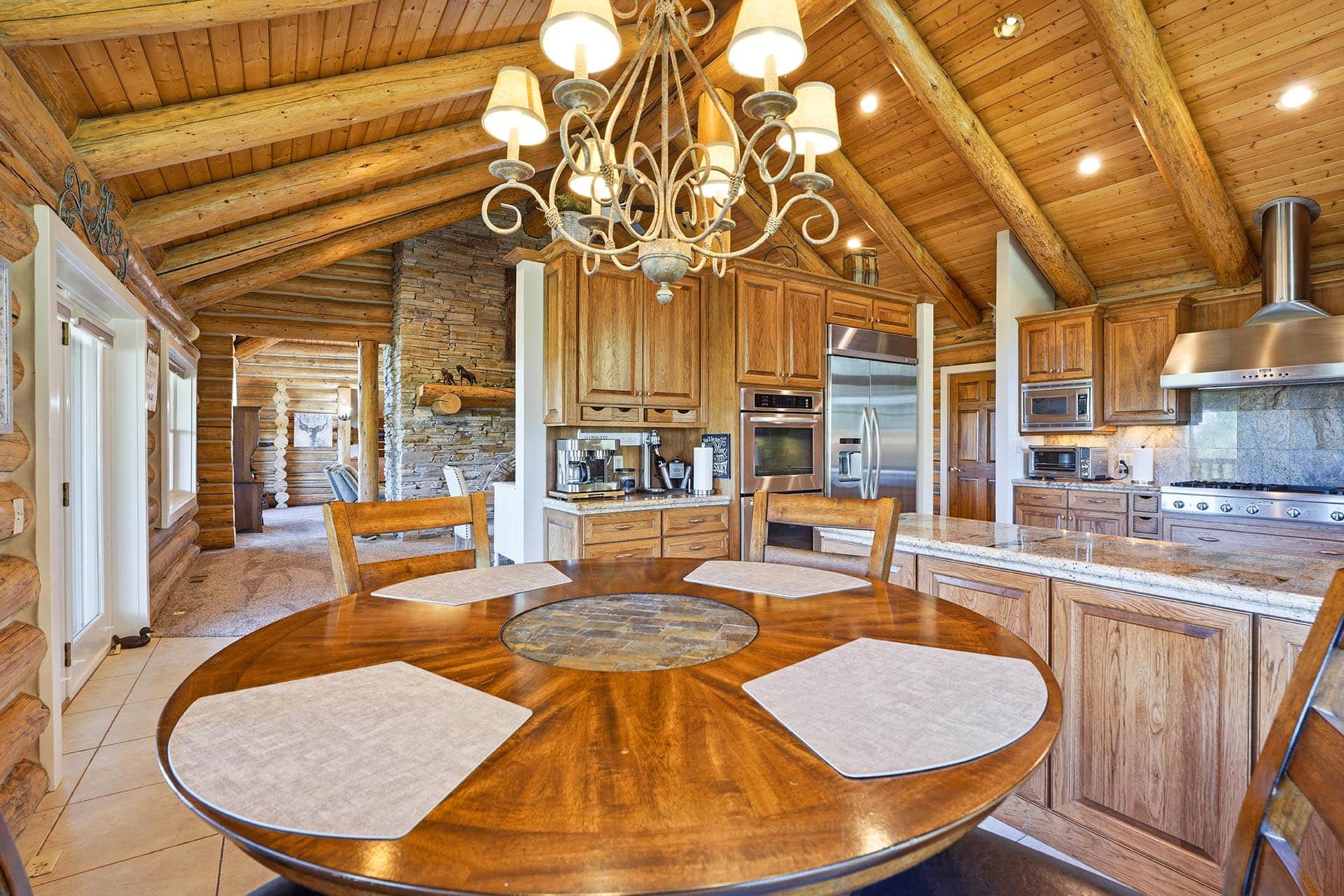 Rustic kitchen with exposed log beams, stone chimney, and wooden cabinets; round dining table in foreground with placemats.