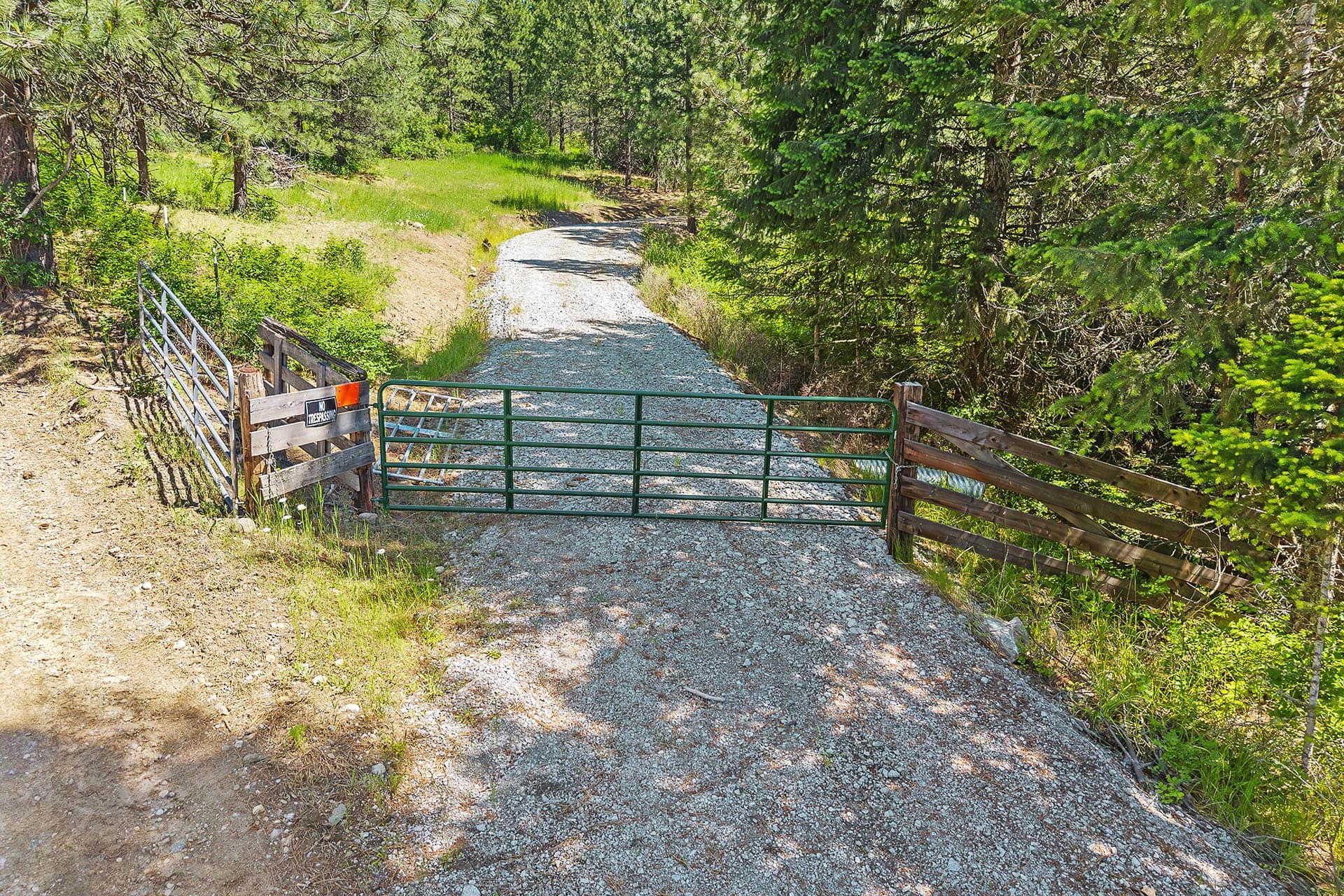 Gravel road blocked by a green metal gate and wooden fence in a forested area; 'No Trespassing' sign visible on the left.