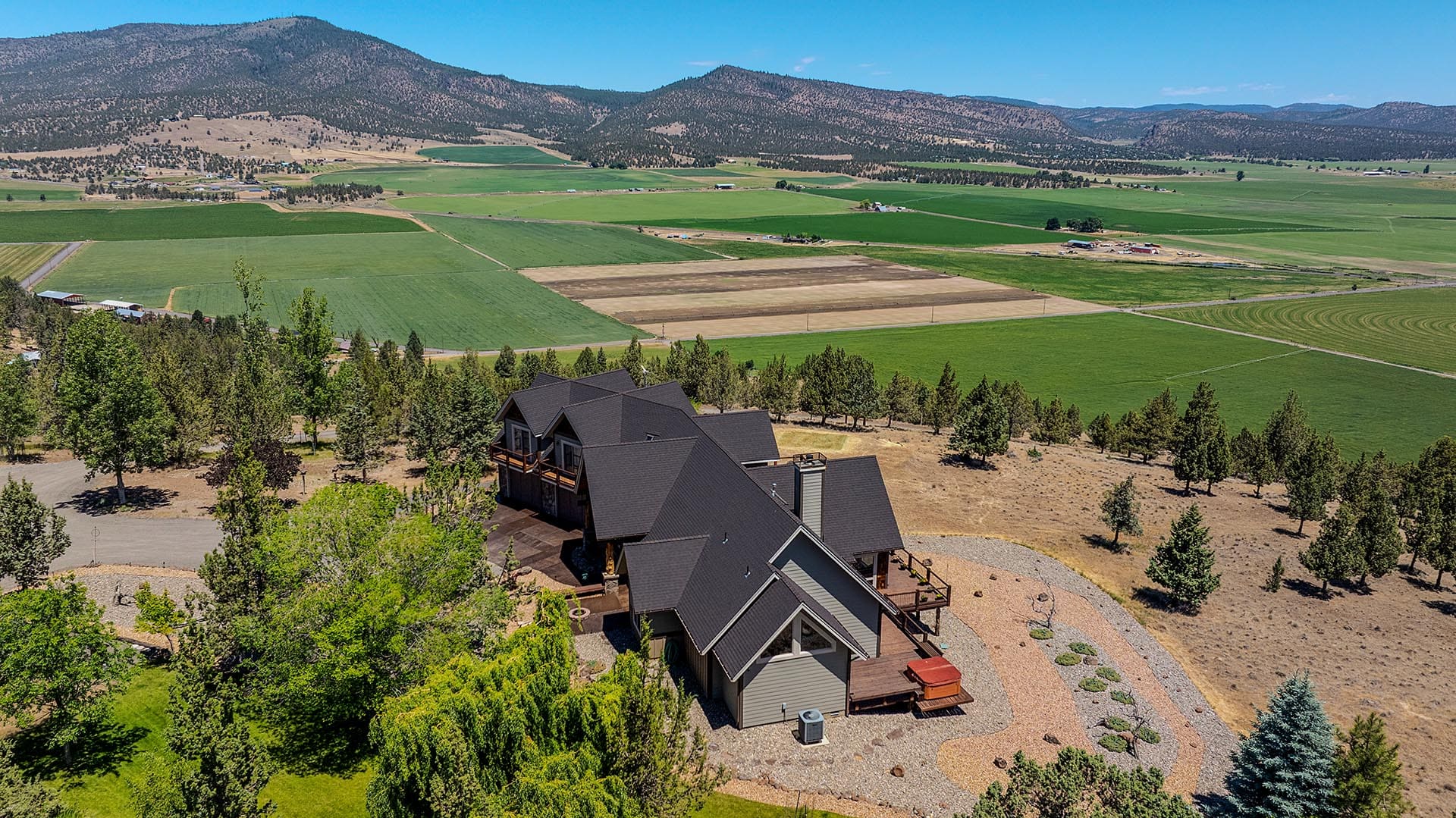 A modern multi-gabled house sits on a hill overlooking patchwork farmland and distant mountains under a bright blue sky.