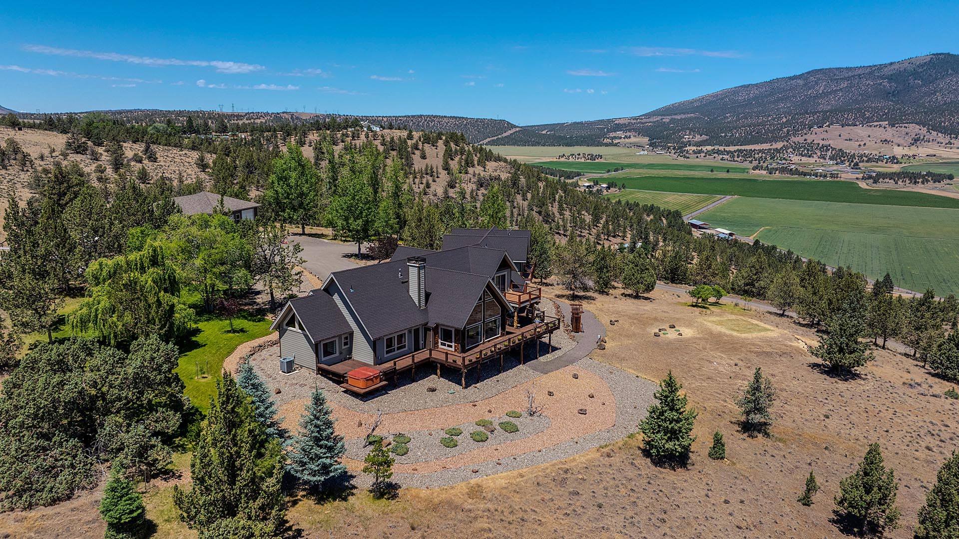 Aerial view of a modern house on a forested hillside with a wooden deck, overlooking green fields and distant mountains.