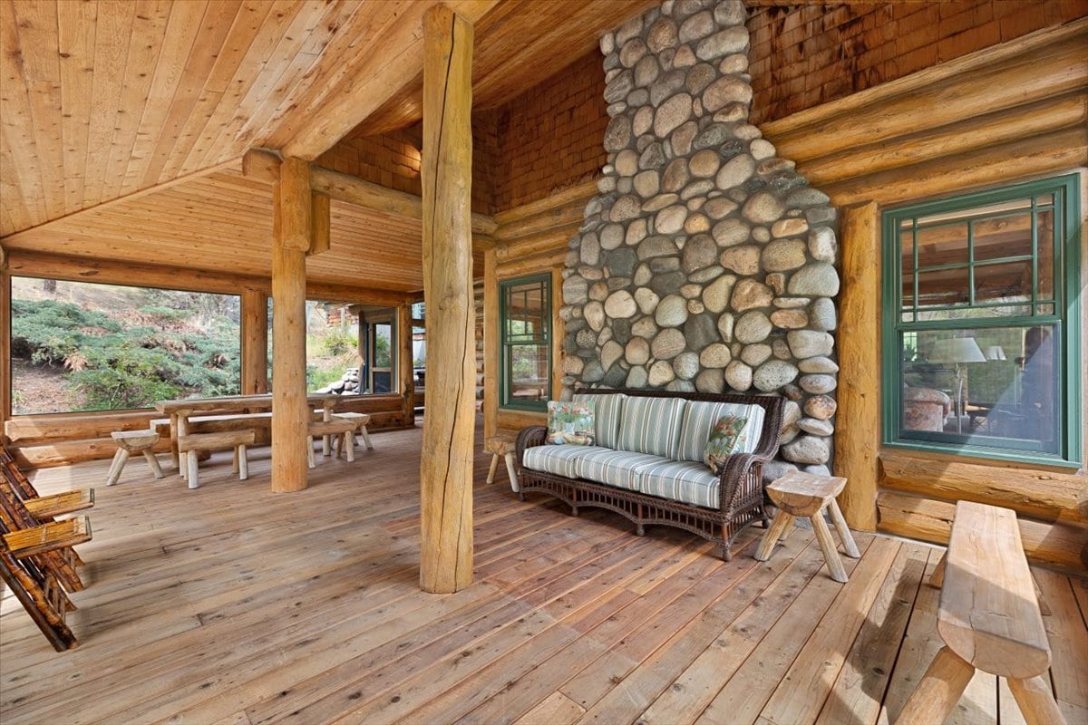 Rustic log cabin porch with a large stone chimney, timber posts, and a striped cushioned wicker sofa.