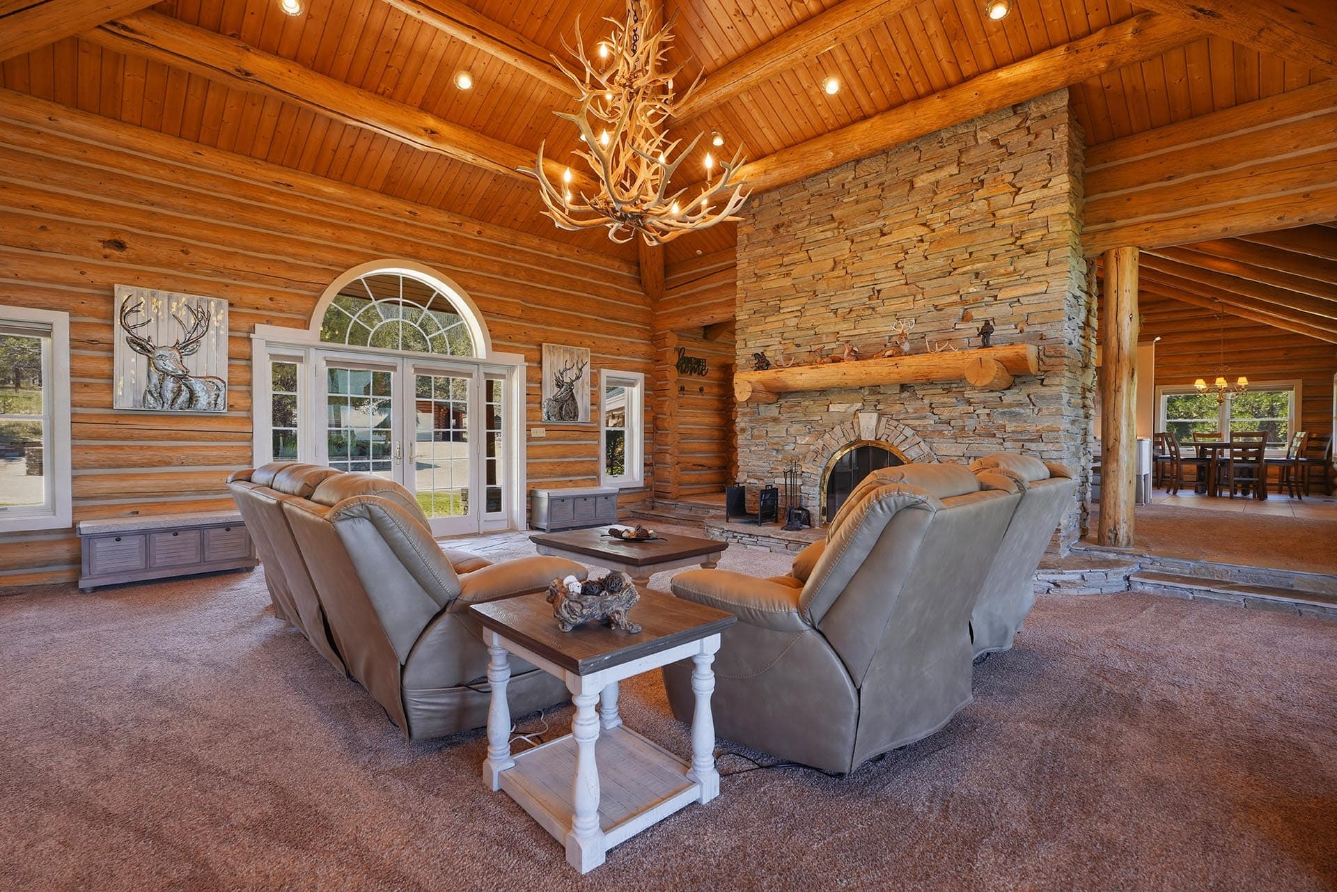 Cozy log cabin living room with a large antler chandelier, stone fireplace, and leather recliners.