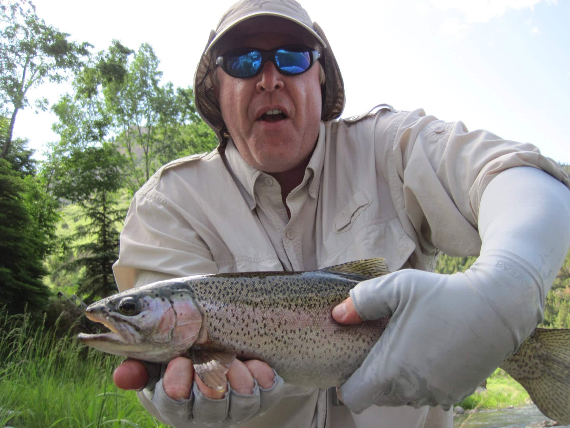 Man in a beige shirt and cap holding a large rainbow trout outdoors by a grassy riverbank with trees behind him.
