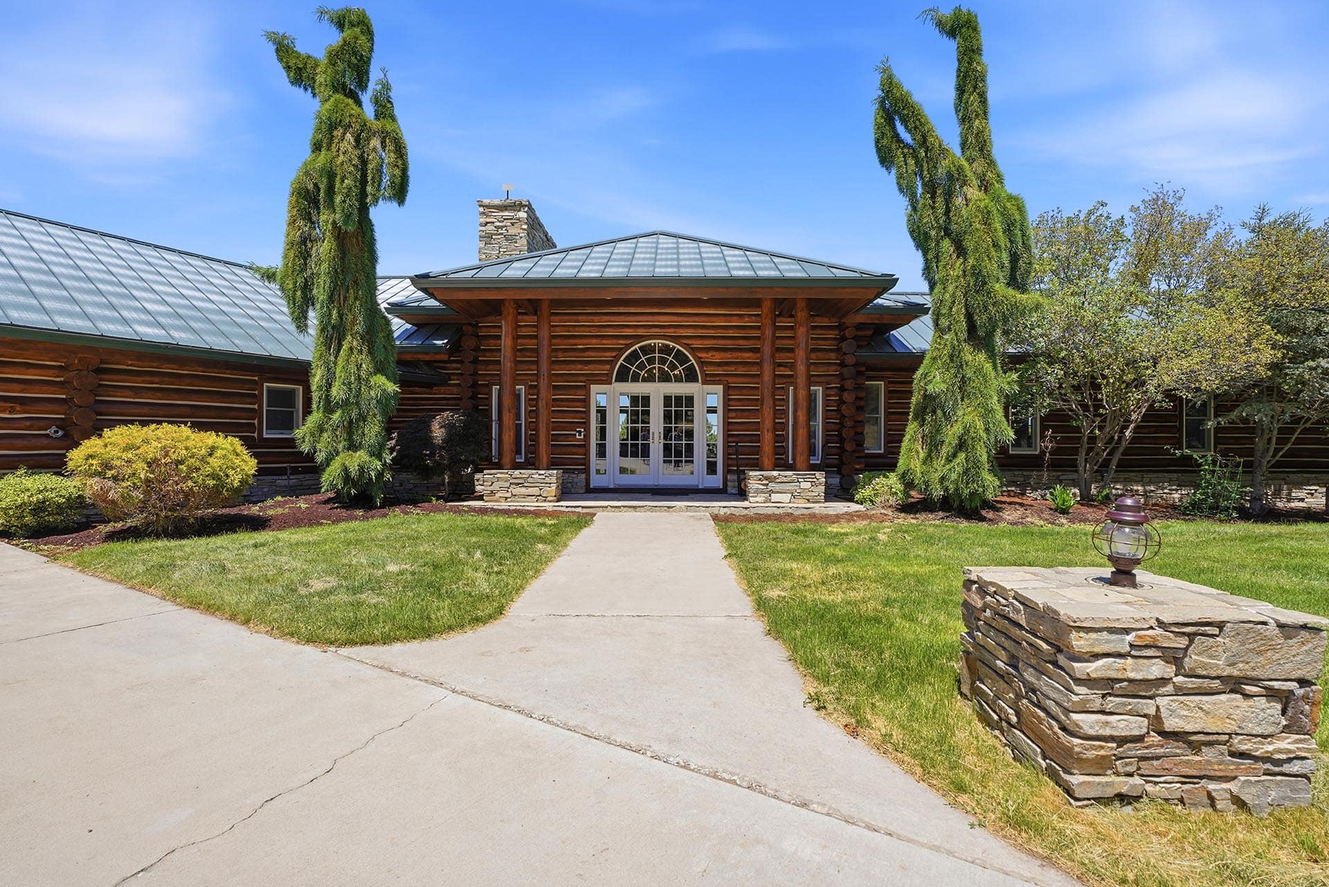 Log cabin-style building with a central arched glass door, stone pillars, and a metal roof, surrounded by manicured landscaping and tall evergreens.