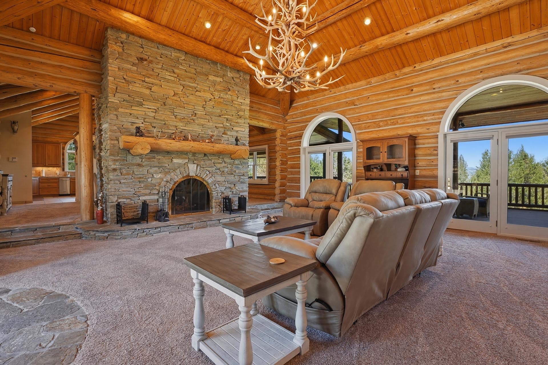 Rustic living room in a log cabin with a stone fireplace, antler chandelier, and leather recliners facing large arched windows.
