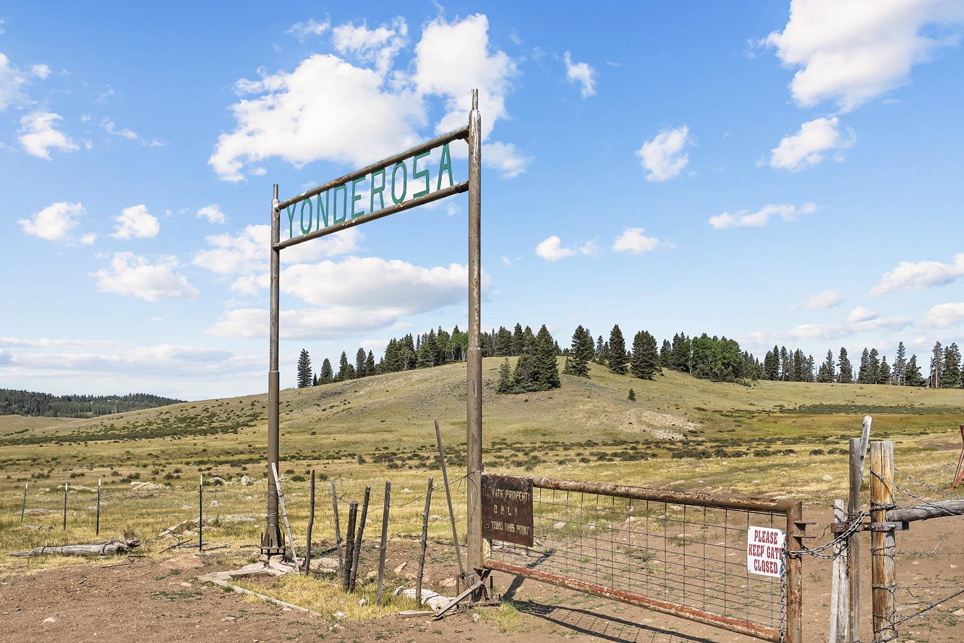 Wooden gate in a rural field with a weathered sign reading 'Yonderosa' overhead; open pasture and pine trees under a blue sky with scattered clouds.