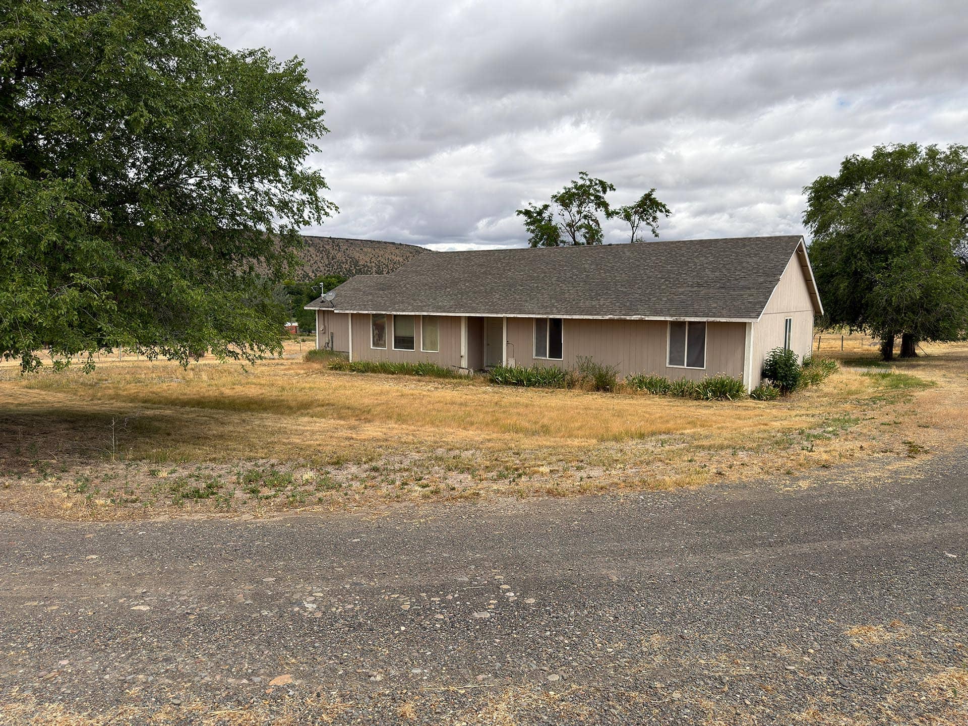 Single-story beige house with gray roof in a dry, rural yard, large tree to the left and cloudy sky above.