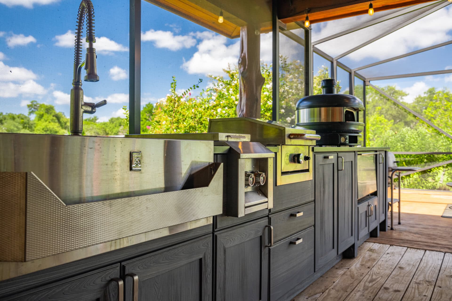Outdoor stainless steel BBQ grill setup with sink, smokers, and storage cabinets on a wooden deck under a covered patio.