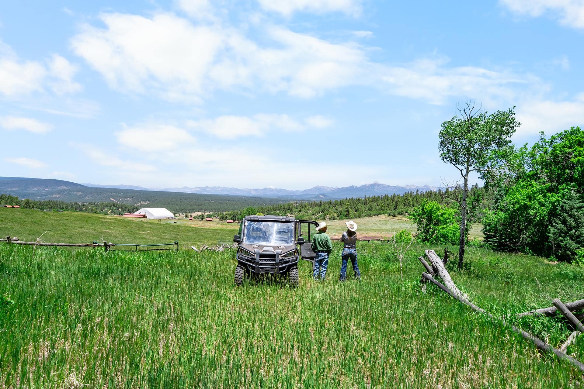 Two people stand beside a dark off-road vehicle in a green field, with distant mountains and a clear blue sky in the background.