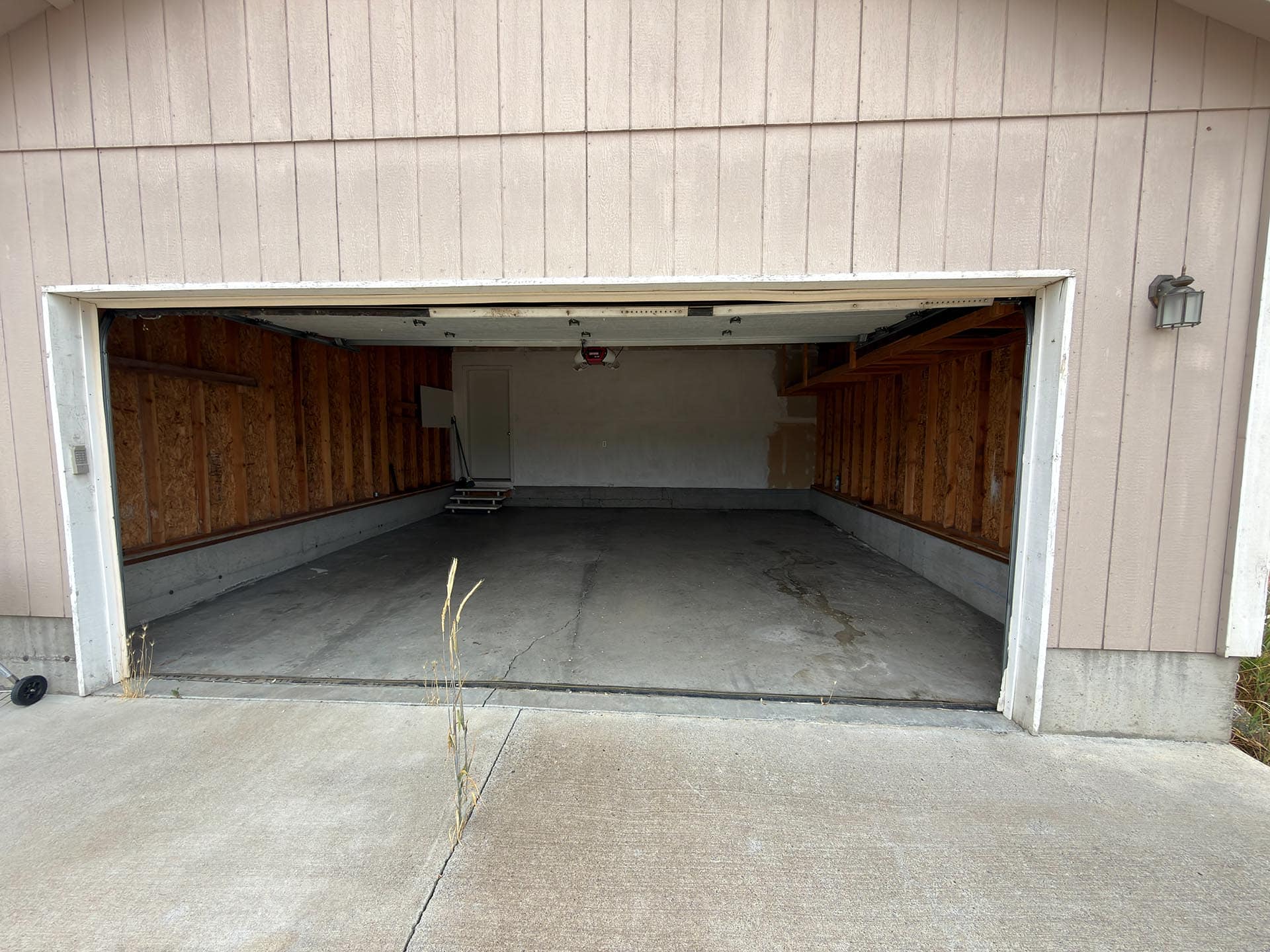 Open garage with unfinished interior: exposed wooden studs, concrete floor, and a doorway at the back; exterior beige siding visible outside.